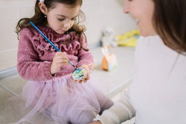 Little girl painting egg using brush on kitchen bar