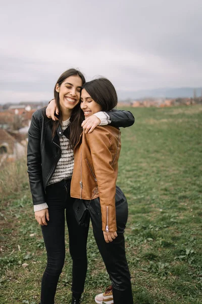 Две смеющиеся девушки обнимаются на поле с видом на город — стоковое фото