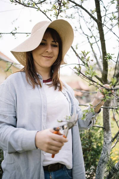 Wanita dengan topi berdiri di taman dan pemangkasan pohon — Stok Foto