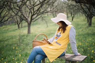 Meyve bahçesinde bankta oturan ve tabletine bakan genç bir kadın.