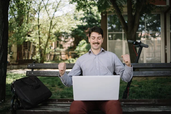 Sonriente hombre sentado en el banco en el parque con el ordenador portátil — Foto de Stock