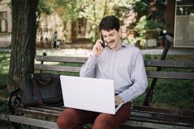 Genç iş adamı dizüstü bilgisayar kullanırken telefonda konuşuyor.
