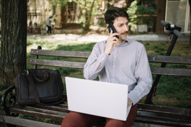 Ciddi bir adam dizüstü bilgisayarla bankta otururken telefonla konuşuyor.