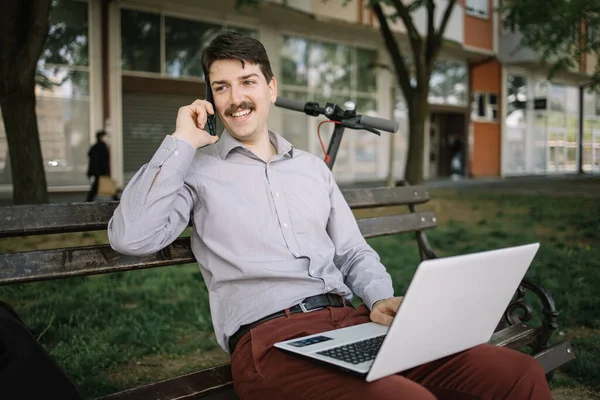 Tipo sosteniendo el teléfono mientras está sentado en el banco con el ordenador portátil — Foto de Stock