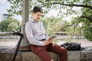 Genç adam çitin üzerinde otururken ve sütuna yaslanırken tableti tutuyor.