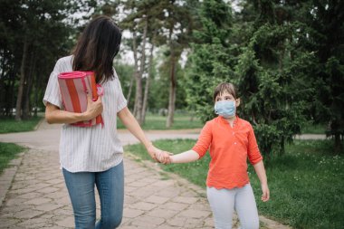 Tıbbi maske takan küçük bir kızla parkta yürüyen esmer bir kadın.