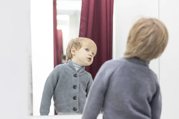 Niño serio: el rubio europeo se prueba la ropa en una tienda en el vestidor y se mira en el espejo. Concepto de compras, estilo y moda . — Foto de Stock