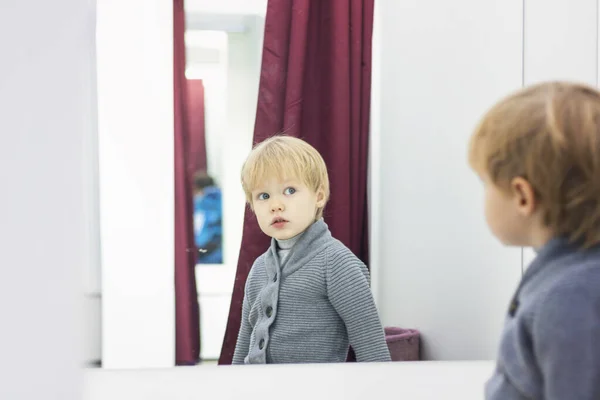 Criança séria - o rapaz louro europeu tenta a roupa em uma loja no camarim e olha para si mesmo no espelho. Conceito de compras, estilo e moda . — Fotografia de Stock