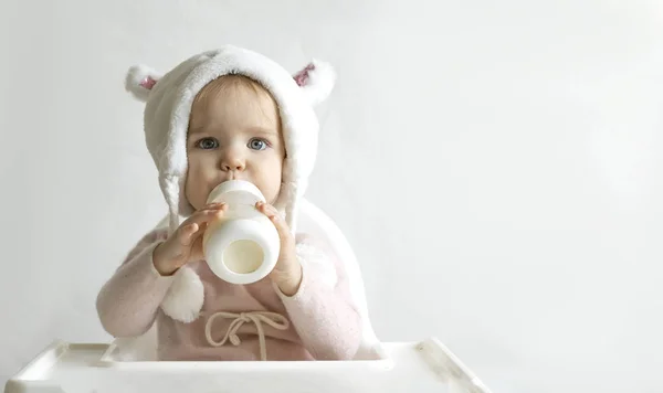 La pequeña niña con un sombrero mullido y cálido bebe leche de una botella mientras está sentada. Retrato de medio cuerpo. Fondo gris blanco . — Foto de Stock