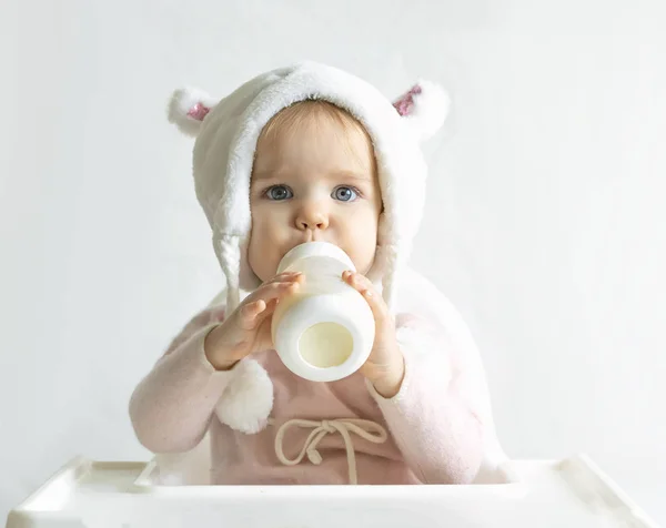 Liten småbarn flicka i en varm fluffig hatt dricker mjölk från en flaska medan du sitter. Halvlångt porträtt. Vit grå bakgrund. — Stockfoto