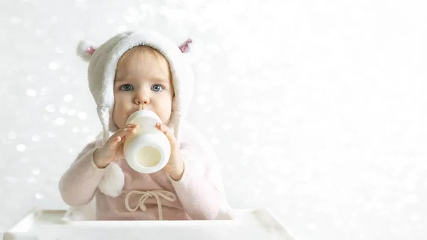 Petite fille tout-petit dans un chapeau moelleux chaud boit du lait à partir d'une bouteille tout en étant assis. Portrait à mi-longueur. Fond gris blanc. Bannière, espace libre, espace de copie — Photo