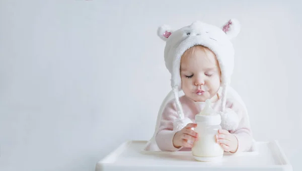 La pequeña niña con un sombrero mullido y cálido bebe leche de una botella mientras está sentada. El niño cerró los ojos de placer. Retrato de medio cuerpo. Fondo gris blanco — Foto de Stock