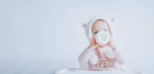 La pequeña niña con un sombrero mullido y cálido bebe leche de una botella mientras está sentada. Retrato de medio cuerpo. Fondo gris blanco — Foto de Stock