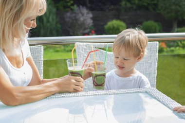 Sarışın bir kadın oğluna sağlıklı bir sebze ve sebze kokteyli veriyor. Çocuklar ve yetişkinler için sağlıklı vejetaryen gıda konsepti bir kır evinin verandasında..