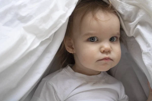 A child of one year old baby looks askance at the camera seriously. Child hid under the blanket. White gray — Stock Photo, Image