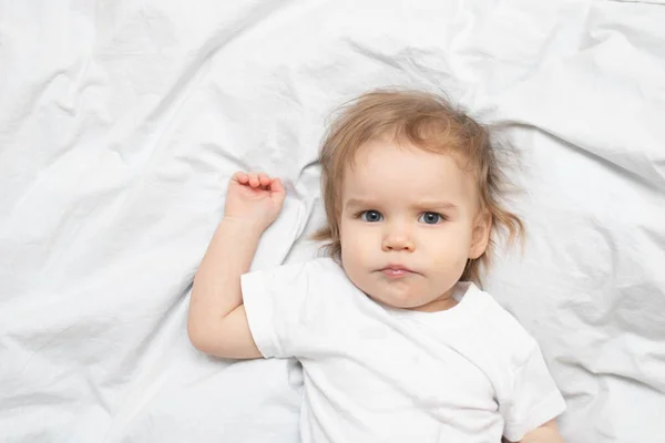 El bebé se acuesta en las sábanas blancas de la cama y mira seriamente a la cámara. Retrato niña un año viejo chico — Foto de Stock