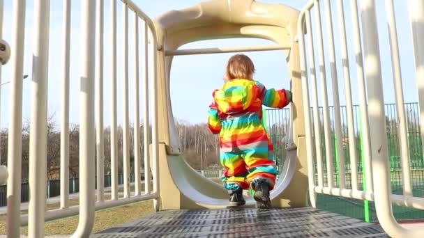 Un niño con ropa de color arco iris cabalga desde un tobogán en un patio de recreo. Primavera, paseos de diversión, paseos y diversión. Niño pequeño rueda cuesta abajo hacia adelante con sus piernas tumbadas sobre su estómago . — Vídeos de Stock