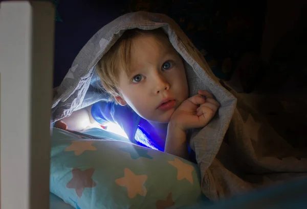 Menino de 3-5 anos jogando no telefone inteligente na cama sob a cama cobre à noite. Pessoas reais — Fotografia de Stock