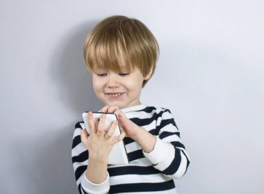 Küçük Avrupalı sarışın çocuk oynuyor, akıllı telefona bakıyor ve gülümseyen kahkahalar atıyor. Uzak Ulak ve video iletişimi ve eğlenceli bir oyun. Gri arkaplan.