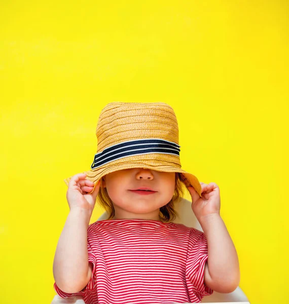 A menina da criança esconde a cara com um chapéu de palha. A criança puxou um chapéu na cabeça sobre um fundo amarelo. Copiar espaço e lugar para texto . — Fotografia de Stock