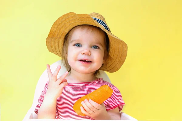 Una niña con sombrero de paja sostiene una zanahoria. El niño muestra tres dedos. El concepto de vitaminas en las zanahorias y el verano jugoso. —  Fotos de Stock