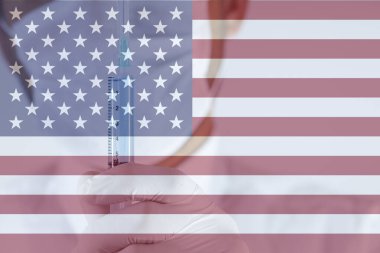 Doktor eldiveninde mavi sıvı olan şırıngayı tutuyor. Yakın plan. Şırınga odaklandı. Doktorun beyaz bir kıyafeti var. Arka planda Amerika Birleşik Devletleri bayrağı.