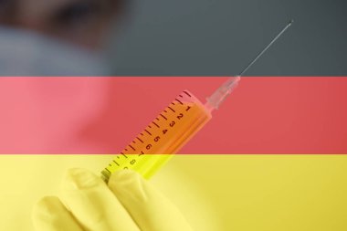 Doktor, eldiveninde sarı sıvı bulunan şırıngayı tutuyor. Yakın plan. Şırınga odaklandı. Doktorun beyaz bir kıyafeti var. Arka planda Almanya bayrağı.