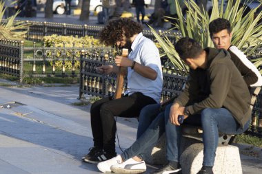 İstanbul, Türkiye - Kasım-07.2019: Sokaktaki kıvırcık saçlı genç adam keman enstrümanıyla şarkı söylüyor. Bankta oturuyor. İnsanlar para verir..