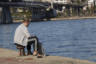 İstanbul, Türkiye - Kasım-02: 2019: Eşofman ve yelek giyen adam bir bit üzerinde oturuyor. O, akıllı telefonuyla ilgileniyor. Altın Boynuz 'un kenarında bir fotoğraf çekilmişti..