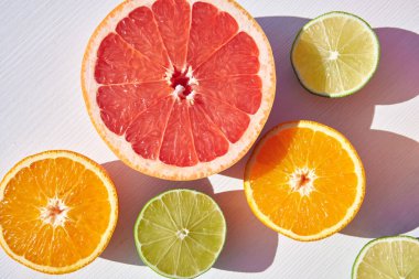 Olgun portakal, greyfurt ve kireç yarımları beyaz ahşap arka planda paketlenmiş, yukarıdan yakın çekim, sert güneş ışığı ve uzun renkli gölgeler..