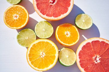 Olgun portakal, greyfurt ve kireç yarımları beyaz ahşap arka planda, yukarıdan yakın çekim, sert güneş ışığı ve uzun renkli gölgeler altında..