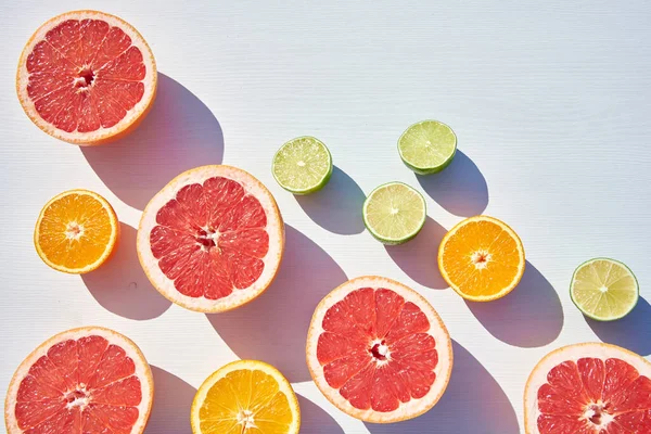 Packed ripe orange, grapefruit and lime halves packed on white wooden background, close-up shot from above in harsh sunlight with long colored shadows.