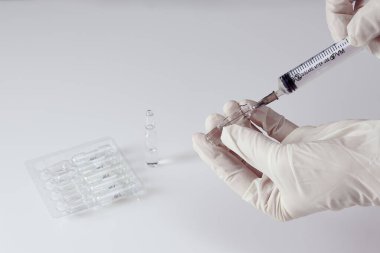 Su toplayan bir su toplayıcısının yanında beyaz akrilik masada ilaç bulunan bir ampul, beyaz tıbbi eldivenli eller şırınganın içine ilaç koyuyor.