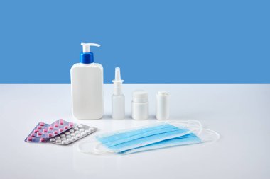 Üzerinde burun spreyi olan beyaz bir antiseptik şişesi beyaz akrilik masada ilaç şişesi ve barut şişesi var. Ön planda su toplayan haplar ve iki mavi ilaç maskesi..