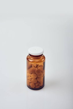 Beyaz plastik kapaklı, yuvarlak sarı tabletlerle dolu, beyaz akrilik masa üzerinde küçük kahverengi bir cam şişenin dikey görüntüsü.