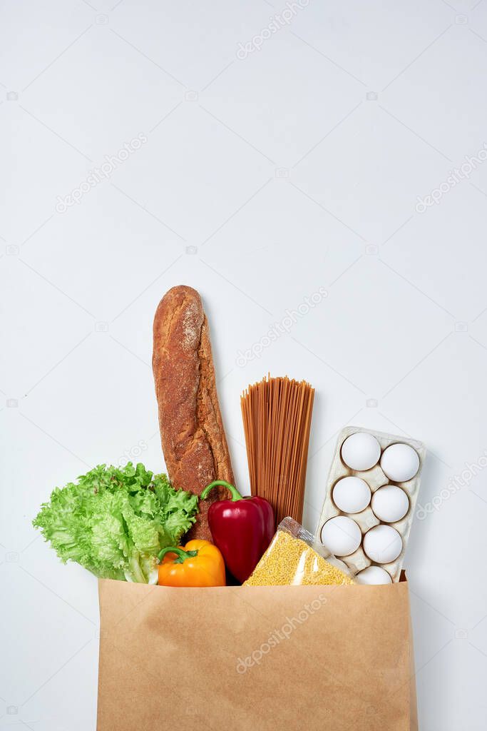 Groceries: bread, buckwheat spaghetti, pack of eggs, lettuce, two bell peppers and pack of groat all in one paper shopping bag on white background