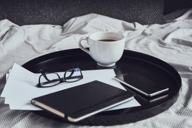 Akıllı telefon, defter, bardaklar, kağıtlar ve siyah yuvarlak tepside bir fincan kahve.