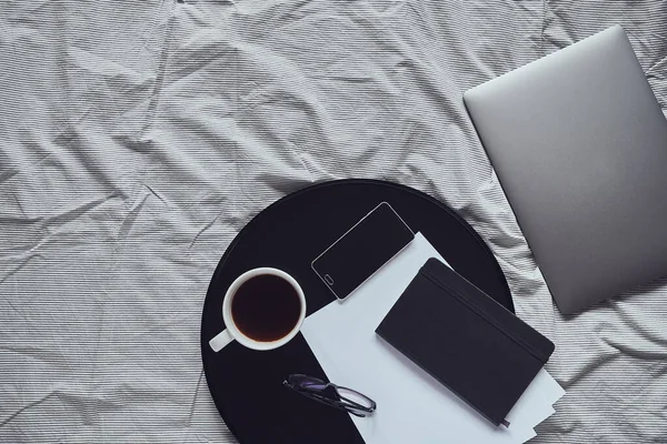 Laptop laying on blanket, next to smartphone, notebook, glasses, sheets of paper and cup of black coffee on black round tray, top view