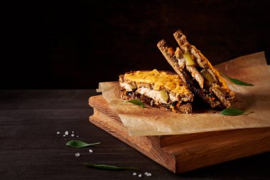 Eritilmiş peynirli tavuk ve karamelize edilmiş soğan sandviçi, biraz yeşillik ve deniz tuzu ile ikiye bölünmüş, geniş ahşap doğrama tahtası üzerinde koyu kahverengi arka planda.