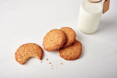 Süt şişesinin yanında beyaz tahta masada etiketli kurabiyeler, bir kurabiye ısırılmış. 45 derece görünüm