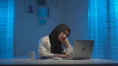 Genç ve güzel Asyalı Müslümanların koyu renk başörtüleri takıp, geceleri evde çalışırken yorgun ve uykulu oldukları modern bir iç tasarım çalışma alanında sıcak mavi ışıkla çalışmalarının odağı.