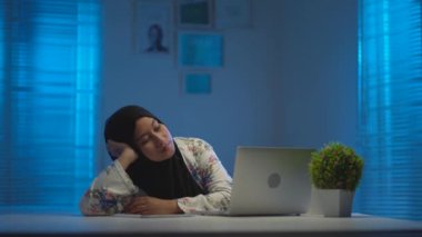 Siyah başörtüsü takan Asyalı güzel Müslümanların yumuşak odağı gece evde çalışırken yorgun ve uykulu oluyorlar. Modern bir iç tasarım çalışma alanında pencerelerden gelen sıcak mavi ışıkla.