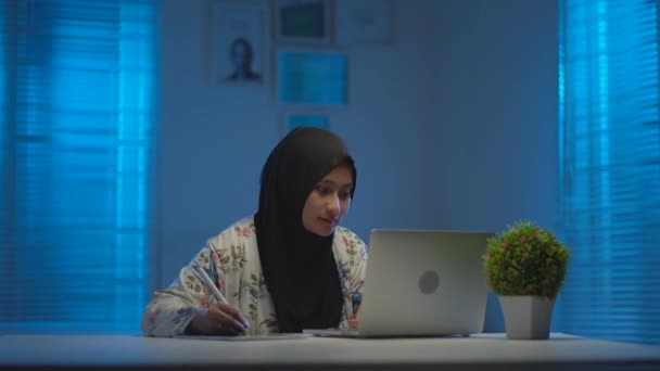 現代的なインテリアデザインのワークスペースで夜に自宅から仕事をするときに楽しむ暗いスカーフを身に着けている若いイスラム教徒のソフトフォーカス窓から暖かい青の光 — ストック動画
