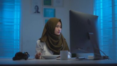Genç Asyalı güzel Müslümanların hafif odak noktası koyu renk başörtüleri takmaları, evde çalışırken yemek yemeleri, modern bir iç tasarım çalışma alanında geceleri sıcak mavi ışıkla çalışmaları.
