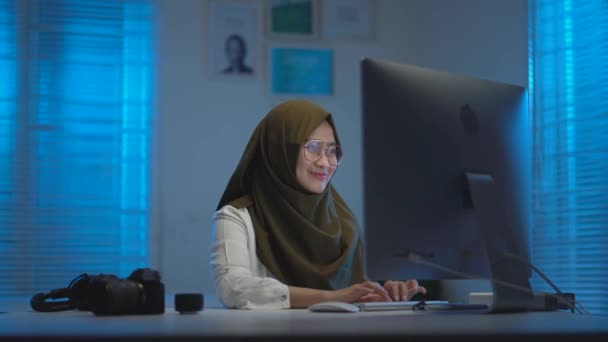 若いのソフトフォーカス暗いスカーフを身に着けている美しいアジアのイスラム教徒は 暖かい青の光と現代的なインテリアデザインのワークスペースで夜に自宅から仕事をしながら とても幸せ — ストック動画
