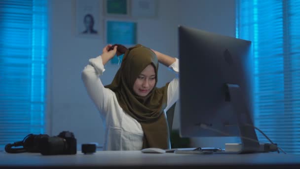 若いのソフトフォーカス自宅から仕事をしながらマスクとヘッドセットを身に着けている暗いスカーフを身に着けている美しいアジアのイスラム教徒は 暖かい青の光と現代的なインテリアデザインのワークスペースで夜のラップトップで作業 — ストック動画