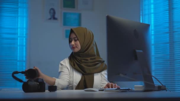 Weicher Fokus junger asiatischer Musliminnen mit dunklen Kopftüchern, die es genießen, von zu Hause aus Musik mit einem Headset zu hören, während sie nachts am Laptop in einem modernen Arbeitsbereich mit warmblauem Licht arbeiten