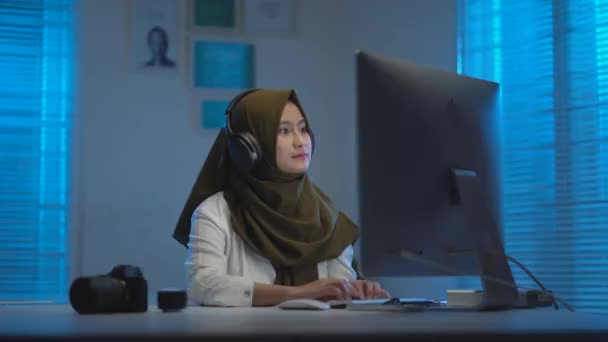 若いのソフトフォーカス暗いスカーフを身に着けている美しいアジアのイスラム教徒は 暖かい青の光と現代的なインテリアデザインのワークスペースで夜のラップトップで働いて 自宅から仕事中にヘッドセットで音楽を聴くのを楽しんで — ストック動画