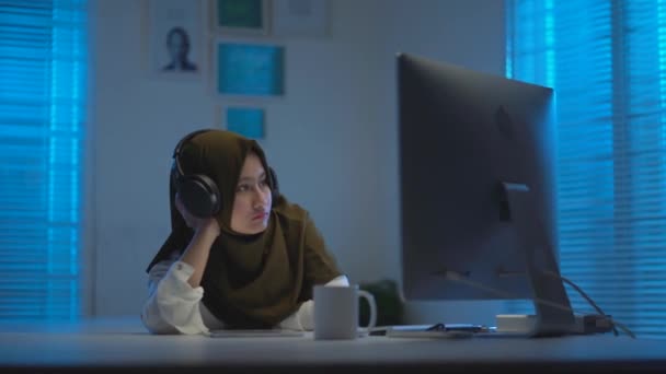 若いのソフトフォーカス暖かい青の光と現代的なインテリアデザインのワークスペースで夜に家から働くときに疲れて眠くなる暗いスカーフを身に着けている美しいアジアのイスラム教徒 — ストック動画