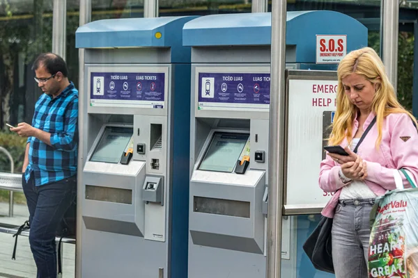 Passagerare som använder sina mobiltelefoner medan de väntar på spårvagnen Luas vid biljettmaskinen på en station i Dublin, Irland — Stockfoto